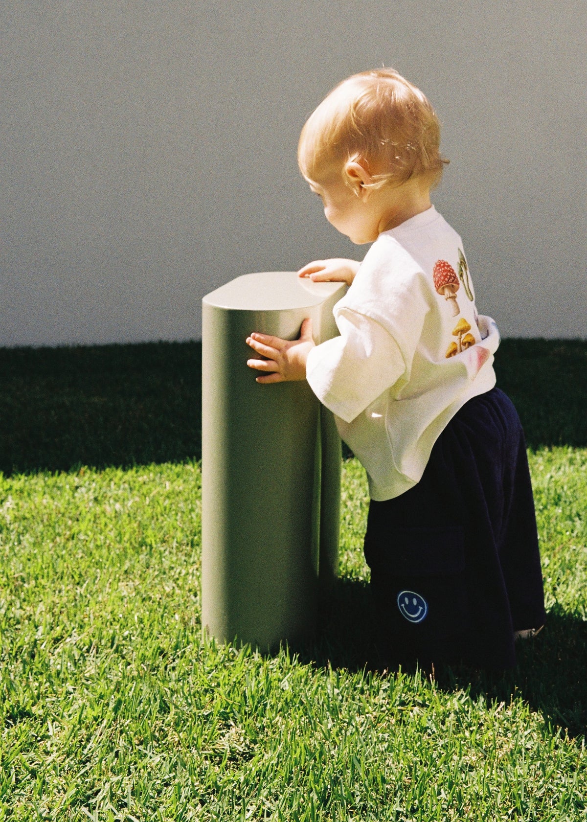 A toddler standing on green grass outdoors wearing the SOZO LAND Little Tee in Mushie Forest, an oversized vanilla cotton T-shirt featuring original colourful mushroom artwork on the front, paired with navy pants.