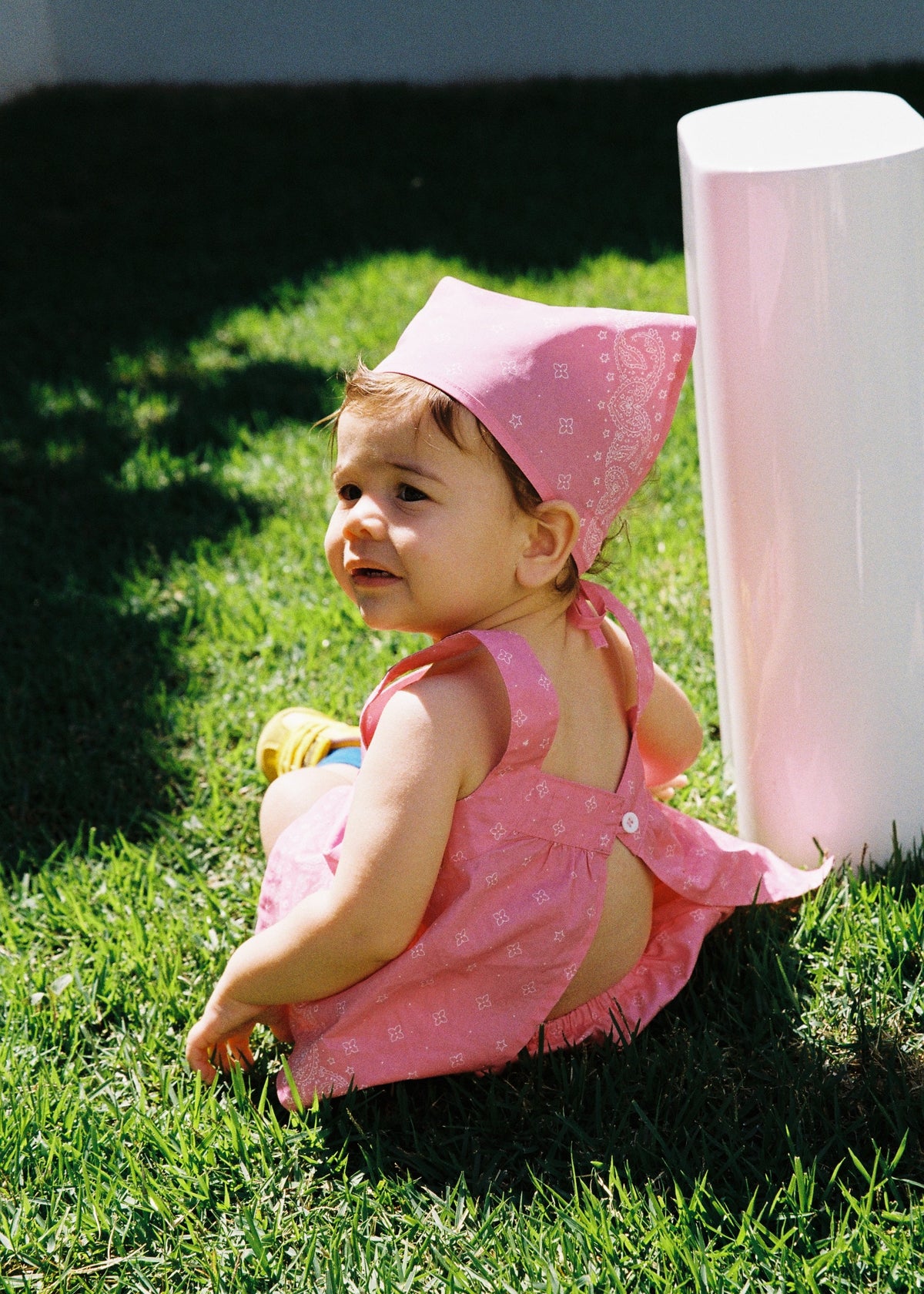 A toddler sitting on green grass outdoors wearing the SOZO LAND Headscarf in Bubblegum pink, featuring our signature Star Paisley print, styled with a matching pink Swing Top and Mini Bloomer.