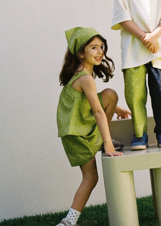 A smiling child wearing the SOZO LAND Headscarf in Lime, featuring our signature Star Paisley print, paired with a matching lime green Swing Top and Mini Skirt, playing outdoors beside a cream-coloured stool.
