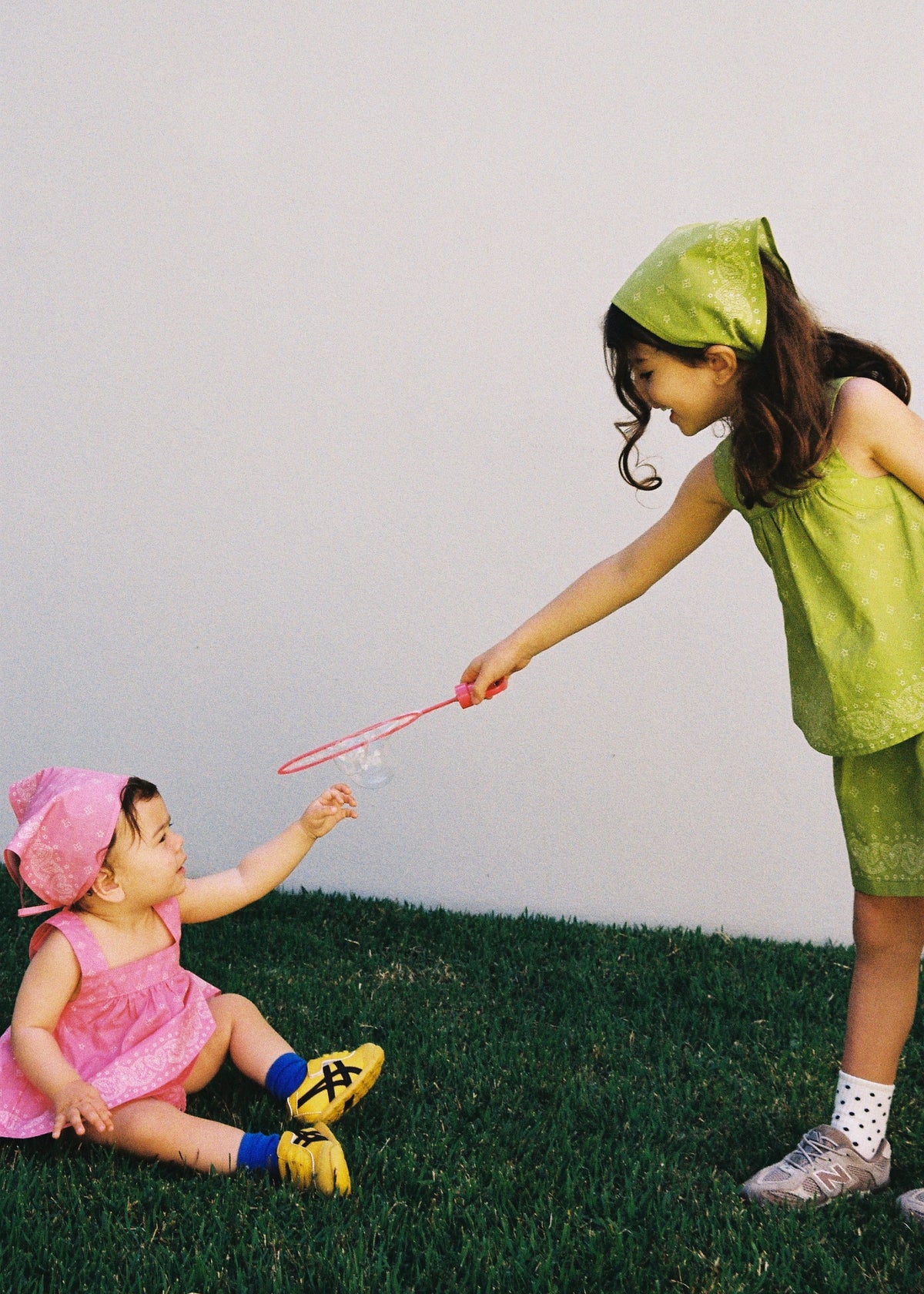 A baby wearing the SOZO LAND Headscarf in Lime with our signature Star Paisley print, being held by a child dressed in pink outdoors in a lush tropical garden.