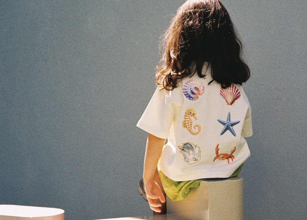 Child photographed from behind wearing the SOZO LAND Little Tee in Sea Dreams, featuring colourful seashell, starfish, seahorse and crab artwork. The oversized white kids tee is styled with lime green clothing against a soft blue-grey backdrop.
