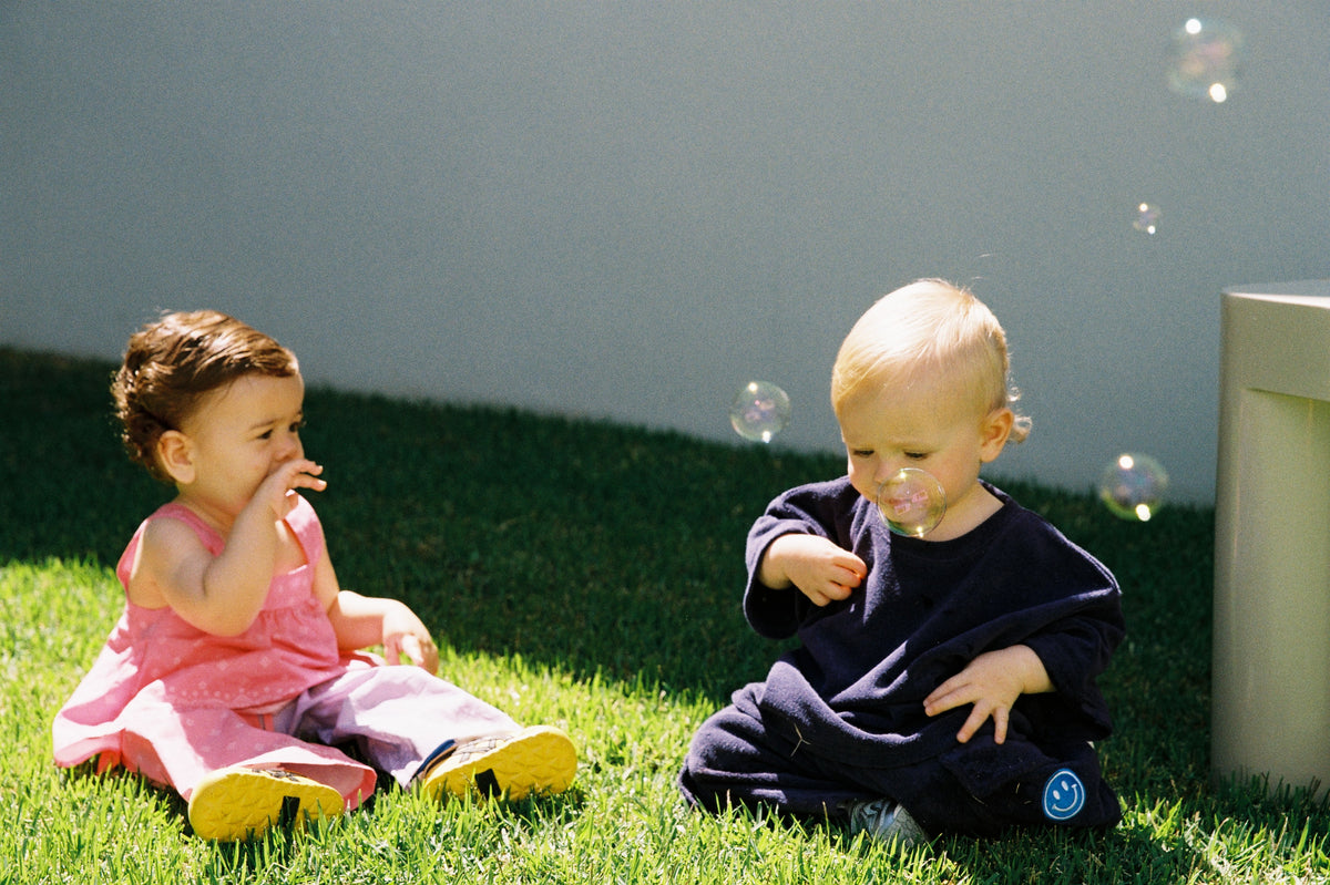 Two toddlers sitting on green grass outdoors, blowing bubbles. One child wears a pink dress and yellow shoes, and the other wears a dark navy outfit.