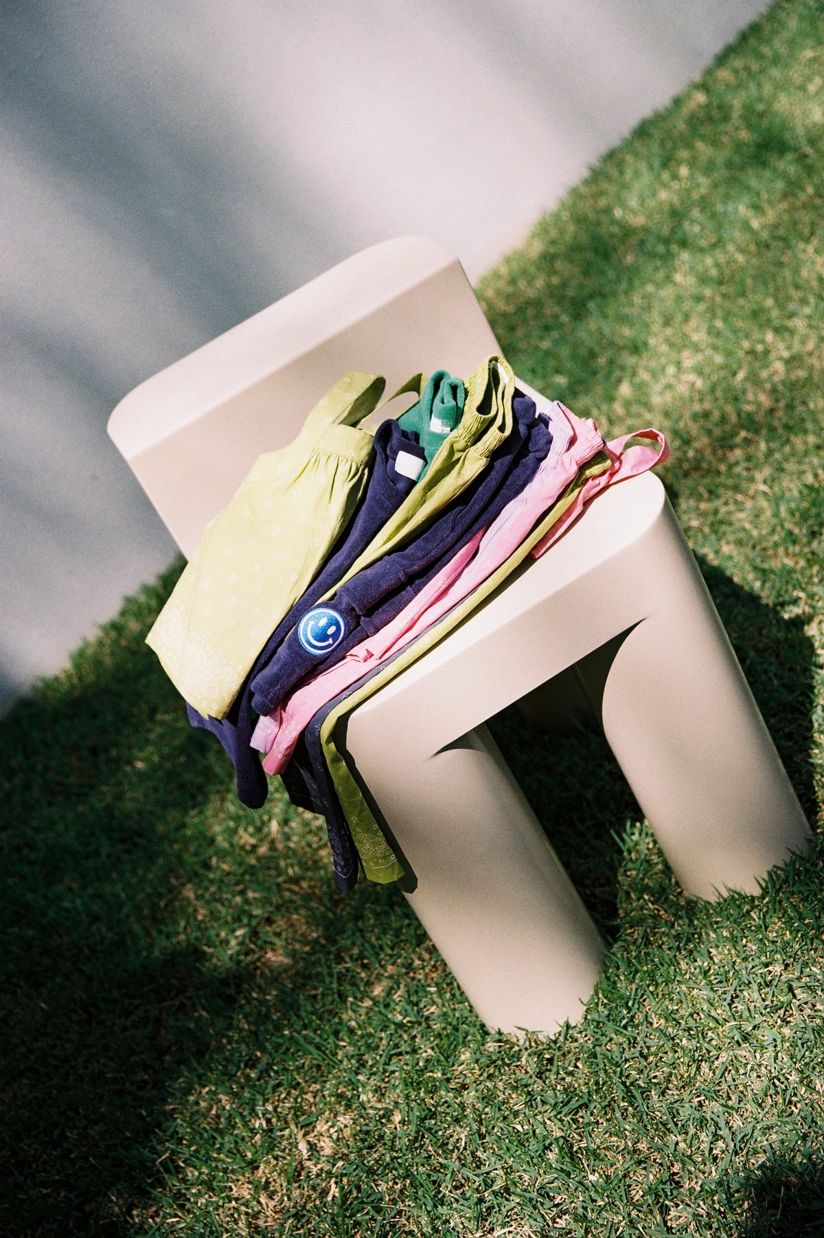 A colourful stack of folded SOZO LAND kids clothing in pink, green, navy and cream, resting on a modern cream chair outdoors on a grassy lawn. Thoughtfully designed Australian childrenswear made for everyday play.
