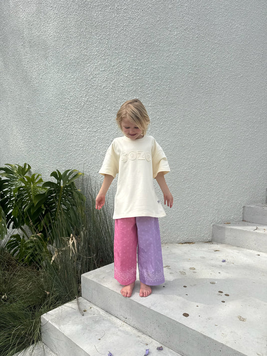 A child standing on outdoor concrete steps wearing the SOZO LAND Tee in Vanilla, an oversized cream kids' T-shirt with a tonal SOZO logo appliqué individually cut and sewn across the front, paired with pink and lilac pants.