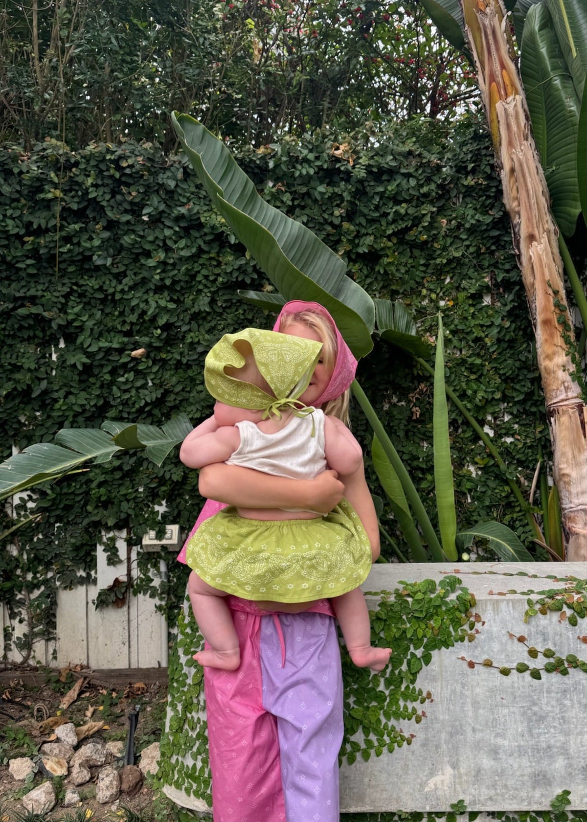 A baby wearing the SOZO LAND Mini Bloomer in Lime with our signature Star Paisley print and matching lime headscarf, being held outdoors in a lush tropical garden.
