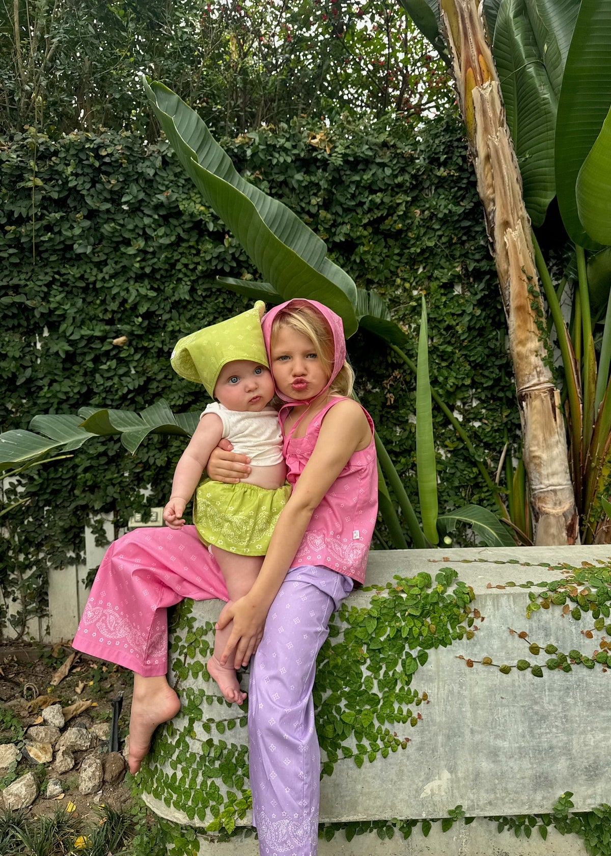Two young children sitting together in a tropical garden, wearing SOZO LAND's Star Paisley headscarves in Bubblegum pink and Lime, styled with colourful matching outfits.