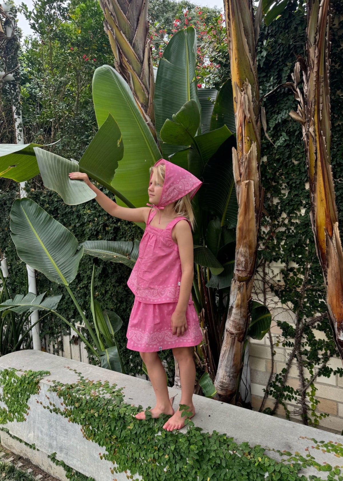 A child exploring a tropical garden wearing the SOZO LAND Mini Skirt in Bubblegum, featuring our signature Star Paisley print, paired with a matching pink Swing Top and Headscarf.