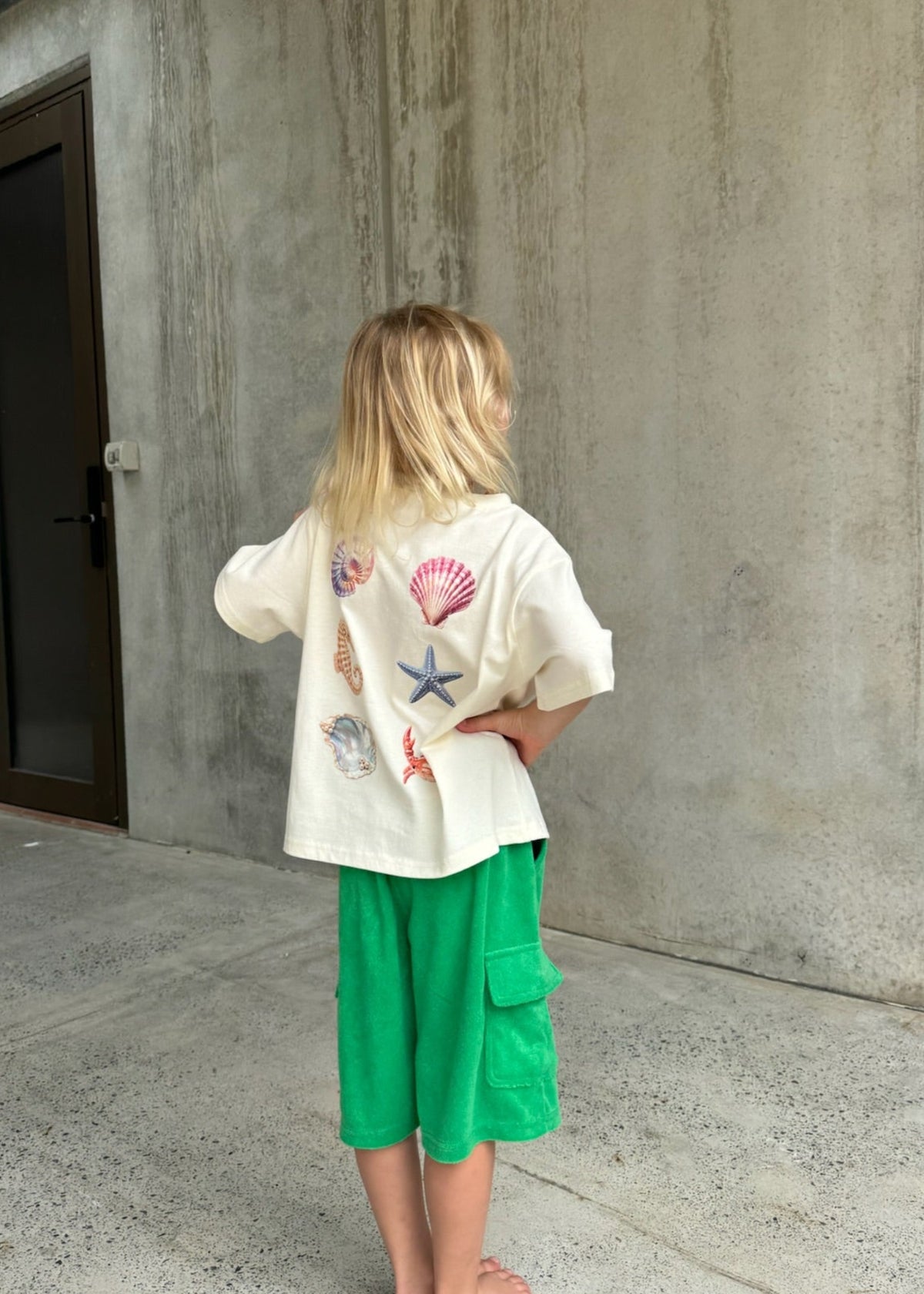 A child wearing the SOZO LAND Little Tee in Sea Dreams, featuring colourful seashell, starfish, seahorse and crab artwork across the back, sitting on a cream play structure outdoors above a baby dressed in green.