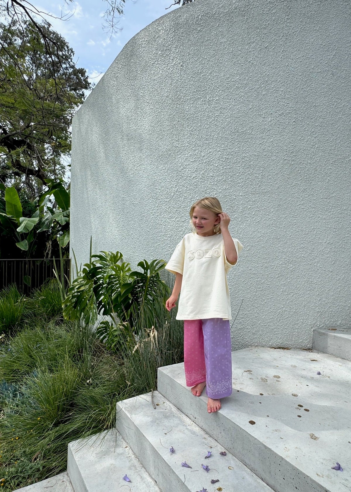 A child standing on outdoor concrete steps wearing the SOZO LAND Play Pant in Bubblegum / Lilac, featuring original Star Paisley print, two-tone pink and lilac colour blocking and a relaxed wide-leg silhouette, paired with an oversized cream tee.