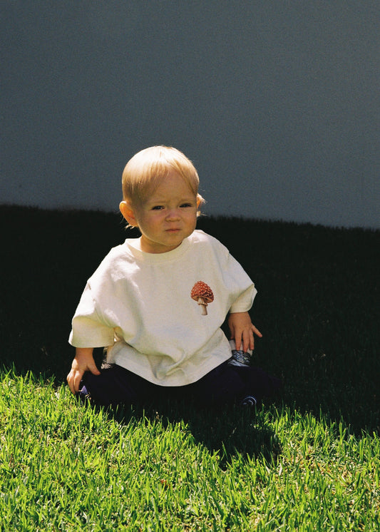 A toddler sitting on green grass outdoors wearing the SOZO LAND Little Tee in Mushie Forest, an oversized vanilla cotton T-shirt featuring original colourful mushroom artwork on the front, paired with navy pants.