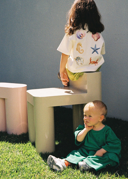 A child wearing the SOZO LAND Little Tee in Sea Dreams, featuring colourful seashell, starfish, seahorse and crab artwork across the back, sitting on a cream play structure outdoors above a baby dressed in green.