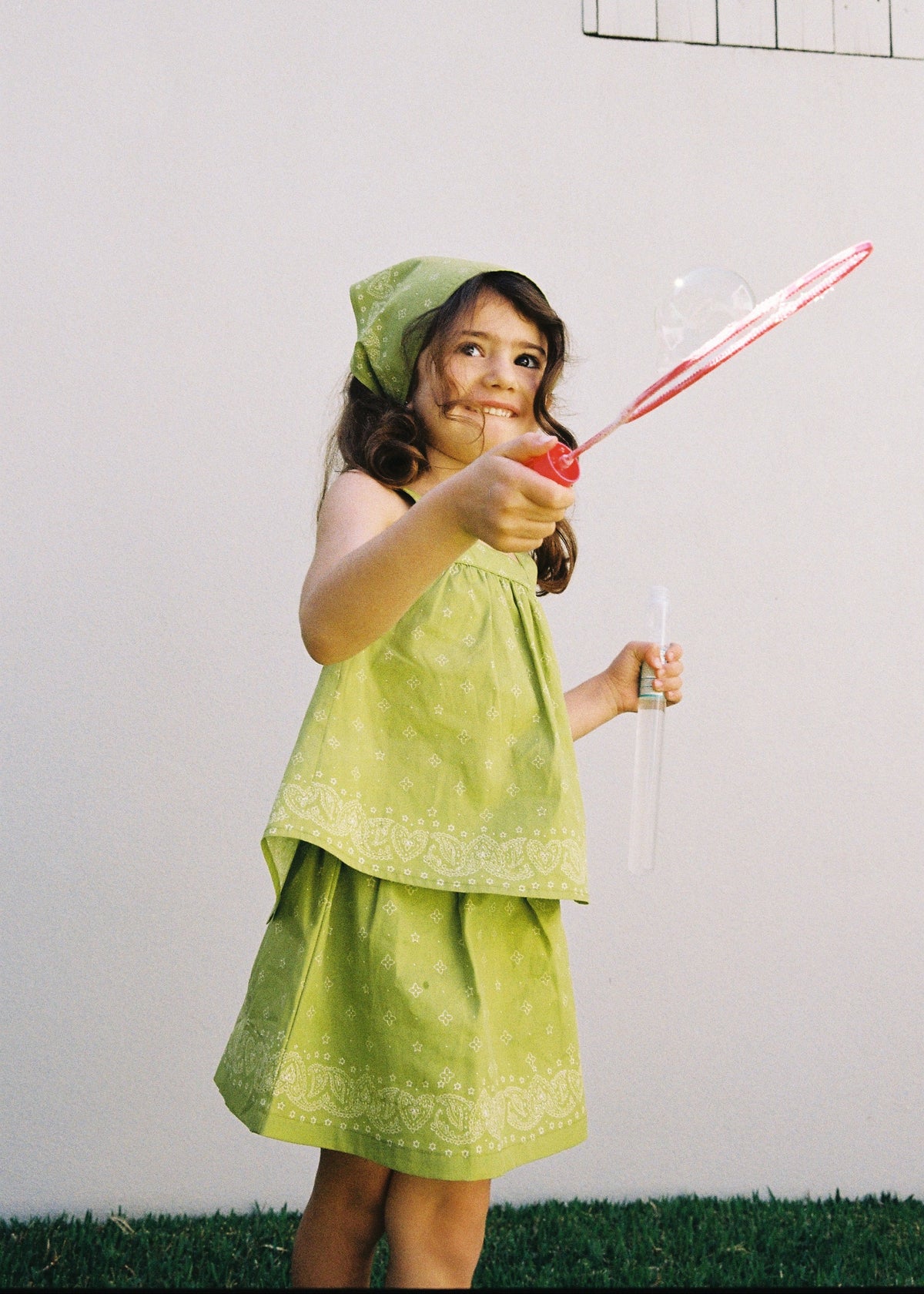 A child playing with a bubble wand outdoors wearing the SOZO LAND Lime Mini Skirt, featuring original Star Paisley print and built-in shorts, paired with a matching Lime Swing Top and Headscarf.
