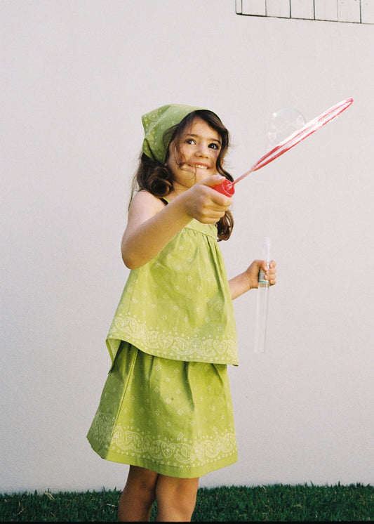 A child playing with a bubble wand outdoors wearing the SOZO LAND Lime Mini Skirt, featuring original Star Paisley print and built-in shorts, paired with a matching Lime Swing Top and Headscarf.