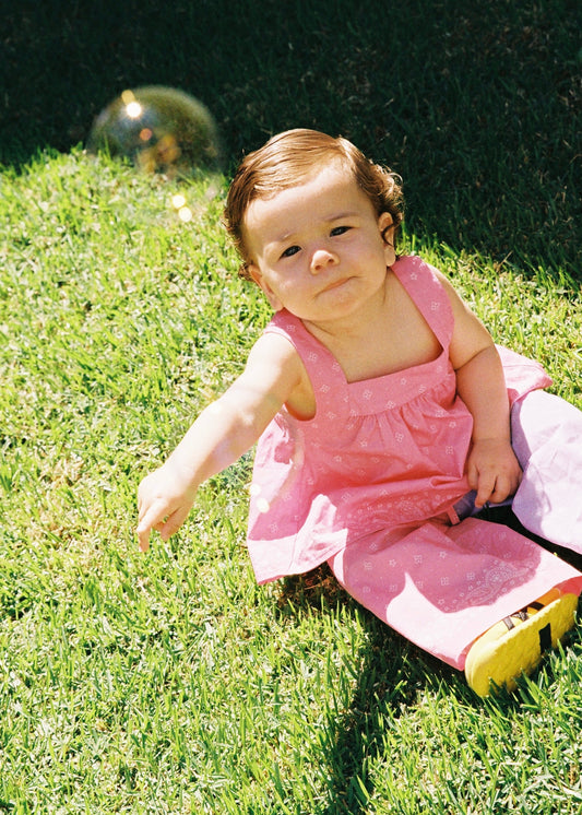 A toddler sitting on green grass outdoors wearing the SOZO LAND Bubblegum Swing Top, featuring original Star Paisley print, shoulder straps and a breezy silhouette, paired with a matching pink Mini Skirt.