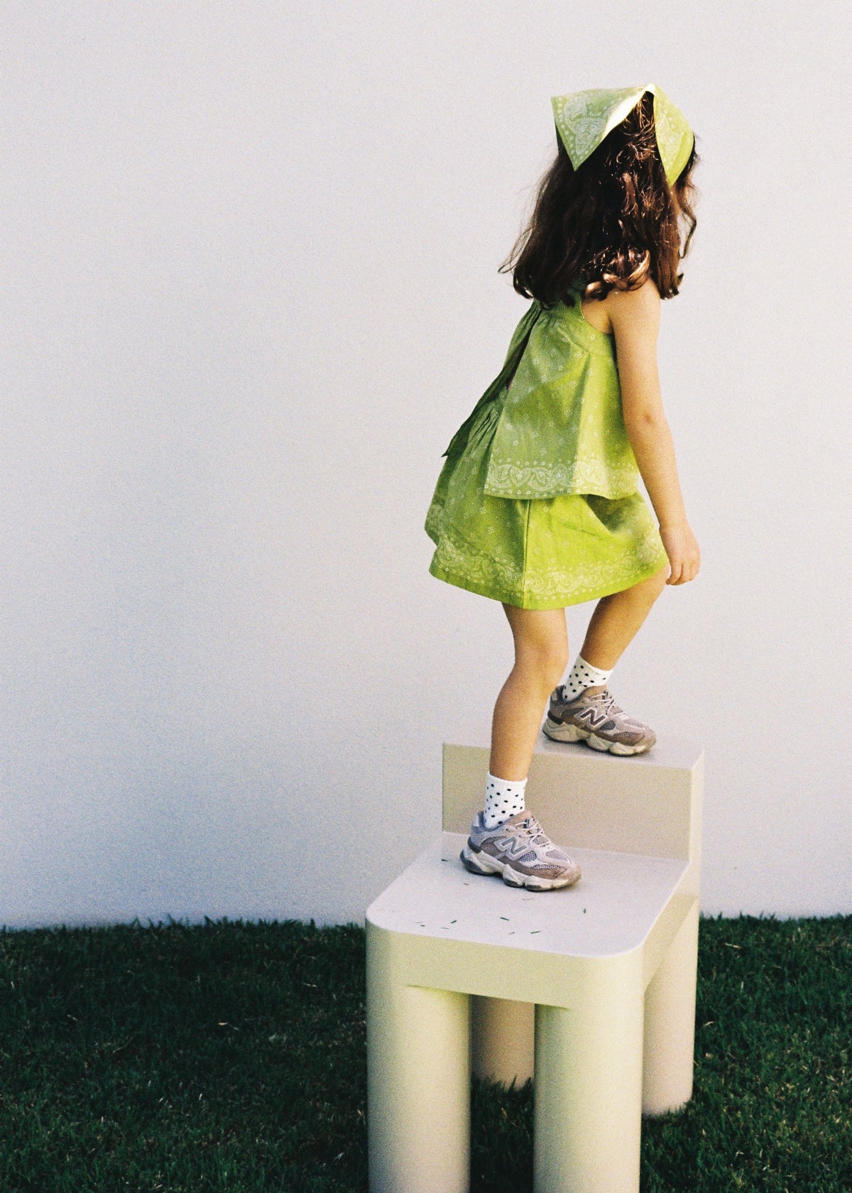 A child standing on a cream chair outdoors wearing the SOZO LAND Lime Swing Top, featuring original Star Paisley print and a breezy open-back silhouette, paired with a matching Lime Mini Skirt and Headscarf.