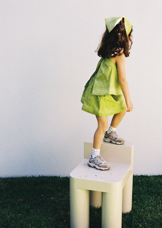 A child standing on a cream chair outdoors wearing the SOZO LAND Lime Swing Top, featuring original Star Paisley print and a breezy open-back silhouette, paired with a matching Lime Mini Skirt and Headscarf.
