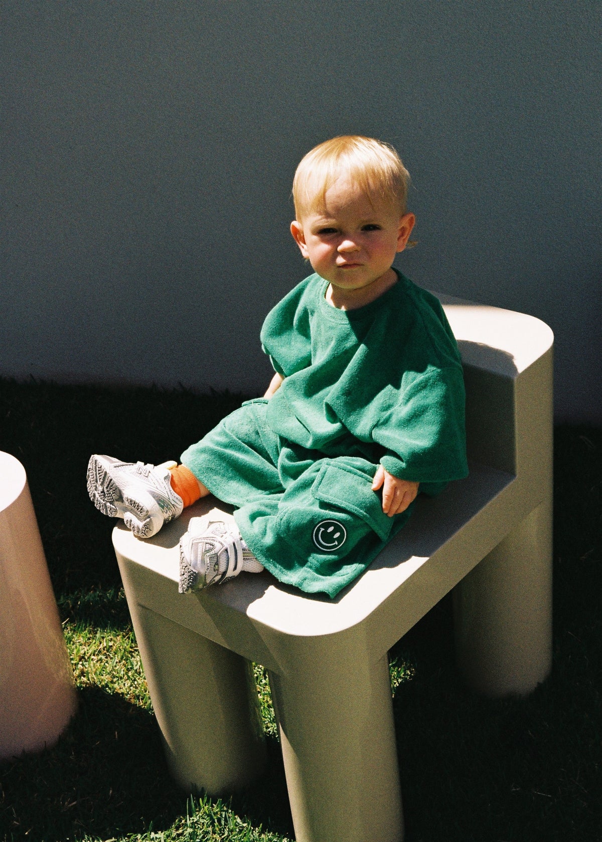 A toddler sitting outdoors on a cream chair wearing the SOZO LAND green Terry Tee and matching three-quarter length Terry Shorts. Made from soft cotton-blend terry, with the signature embroidered SOZO smiley on the shorts.
