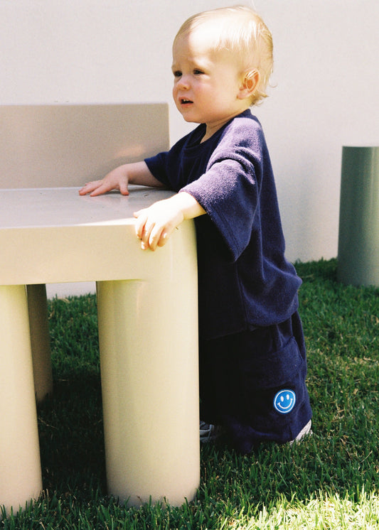 A toddler standing outdoors beside a cream table wearing the SOZO LAND Navy Terry Tee, an oversized kids T-shirt in soft cotton-blend terry, paired with matching navy Terry Shorts featuring an embroidered SOZO smiley.