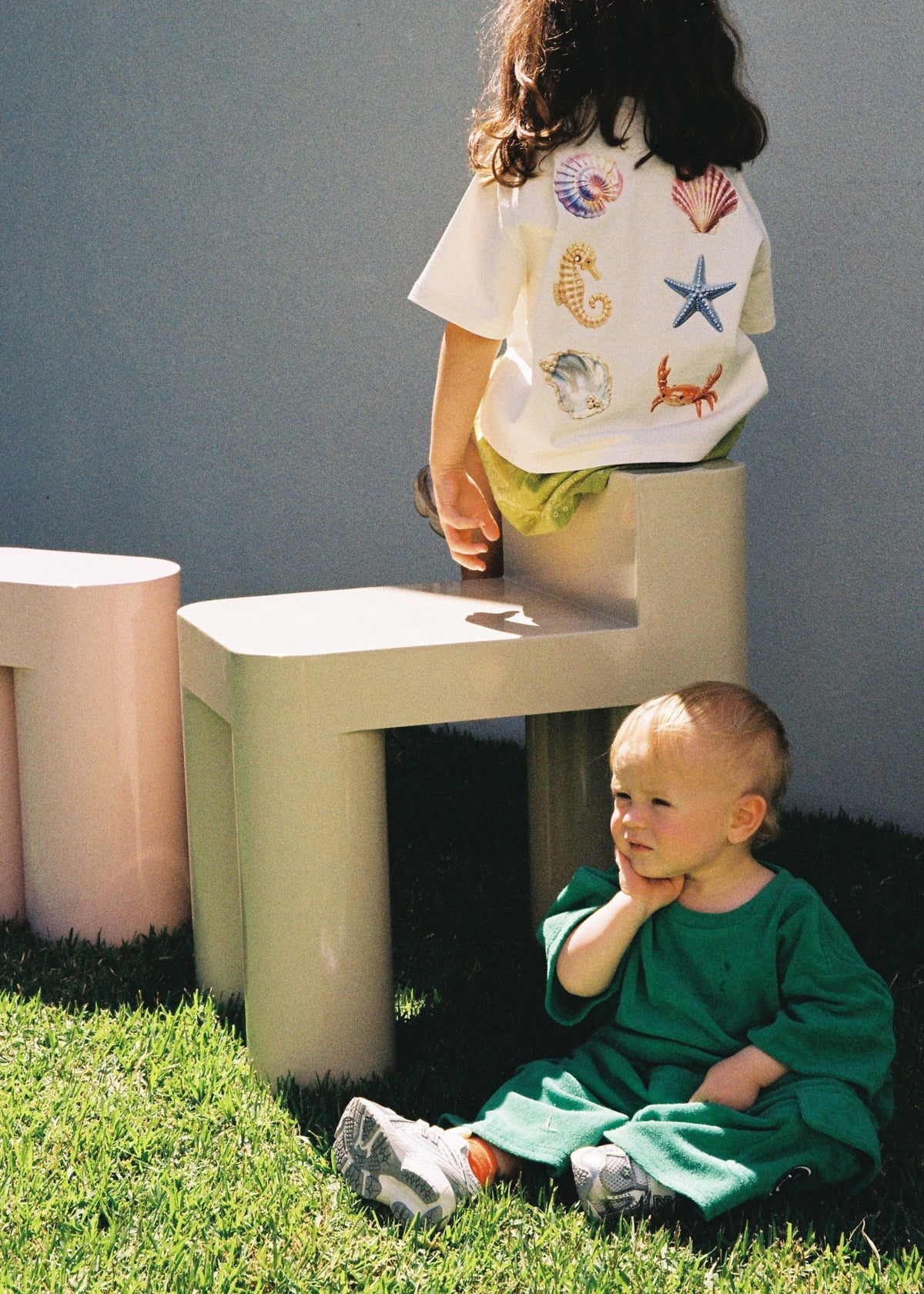 Two children in SOZO LAND clothing playing outdoors on a grassy playground. One wears the Sea Dreams Little Tee with colourful seashell, starfish and seahorse artwork, while the other wears a green outfit.