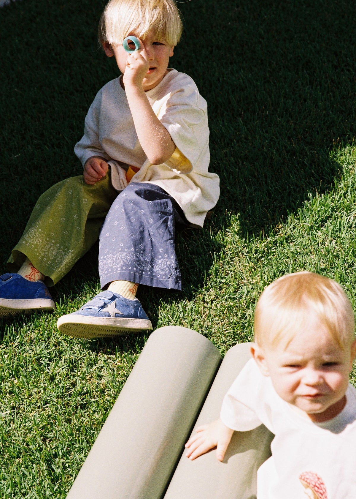 A child sitting on green grass outdoors wearing the SOZO LAND Play Pant in Lime / Navy, featuring original Star Paisley print, a two-tone colour-blocked design and relaxed wide-leg silhouette, paired with a white tee.