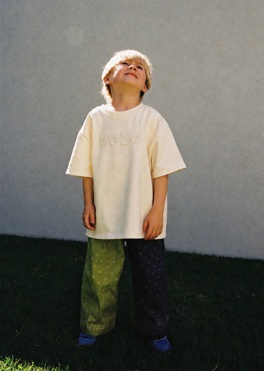 A child standing outdoors on green grass wearing the SOZO LAND Tee in Vanilla, featuring an individually cut and sewn SOZO logo appliqué across the front, an oversized silhouette and colour-blocked lime and navy pants.